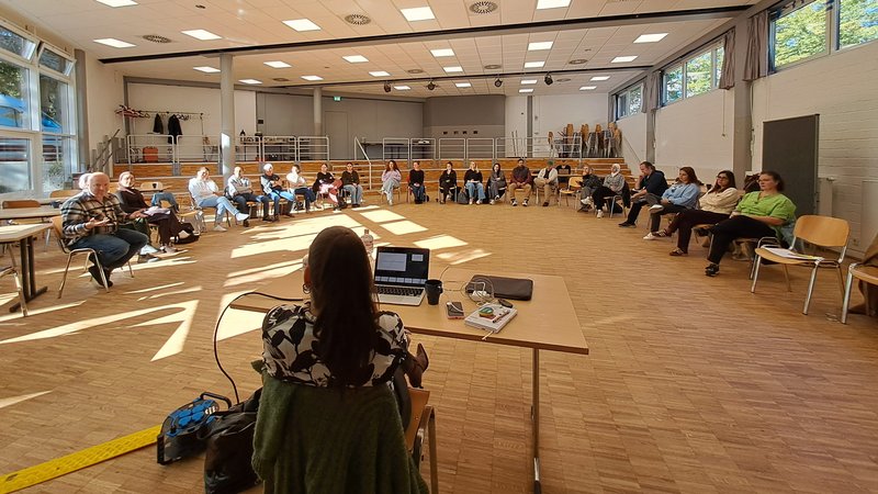 Auf dem Foto ist eine Gruppe von Erwachsenen zu sehen, die an einer Versammlung, Schulung oder einem Workshop in einem großen Raum teilnehmen. Hier sind die Details: Personen und Anordnung: Etwa 20 bis 25 Personen sitzen in einem großen, offenen Raum. Sie sind in einem Halbkreis auf Stühlen angeordnet und schauen in Richtung des vorderen Bereichs. Eine weibliche Person sitzt vorne an einem Tisch mit einem Laptop und ist dem Publikum zugewandt (ihr Rücken ist zur Kamera). Sie scheint die Moderatorin oder Vortragende zu sein. Raum: Es handelt sich um einen großen Mehrzweckraum oder eine Aula mit hellem Holzfußboden. Der Raum hat große Fenster an den Seiten, durch die viel natürliches Licht einfällt. Man sieht Bäume und blauen Himmel draußen. ☀️ Die Decke besteht aus vielen quadratischen Lichtelementen (Rasterdecke). Es gibt eine Bühnentechnik mit Strahlern und einem Beamer/Projektor (der auf eine nicht sichtbare Leinwand projiziert). Im Hintergrund gibt es eine erhöhte Tribüne oder Sitzreihe und gestapelte Stühle, was auf die Nutzung als Versammlungsraum hindeutet. Zusammenfassend zeigt das Bild eine organisierte Veranstaltung in einem großen, hellen Raum.