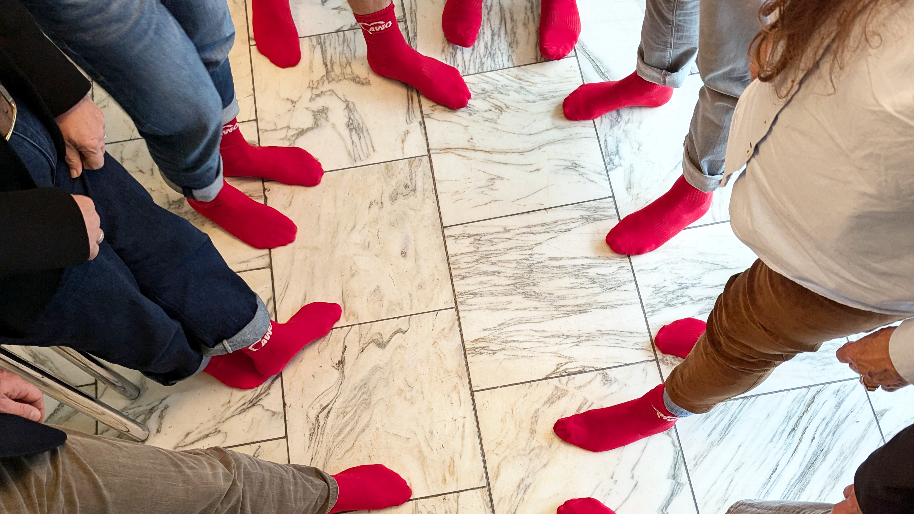 Das Foto zeigt eine Gruppe von Menschen, die in einem Kreis stehen. Sie tragen alle leuchtend rote Socken und sind von der Hüfte abwärts zu sehen. Der Boden besteht aus weiß-marmorierten Fliesen, was einen starken Kontrast zu den roten Socken bildet. 