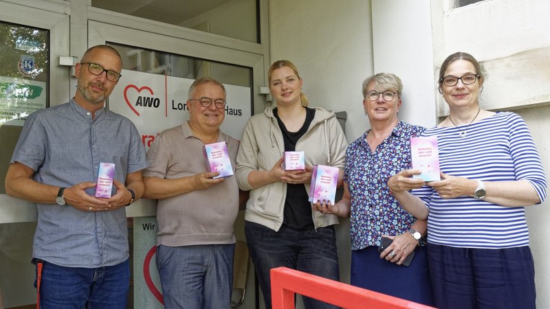 Auf dem Foto sind fünf Personen zu sehen, die vor einem Gebäude stehen. Jede Person hält ein Menstruationsprodukt in der Hand. Neben dem Eingang des Gebäudes ist ein Schild mit dem Logo und dem Namen “AWO” zu sehen, was für “Arbeiterwohlfahrt” steht. Darunter steht “Lore-Agnes-Haus,” was darauf hinweist, dass es sich um eine Einrichtung der AWO namens Lore-Agnes-Haus handelt.