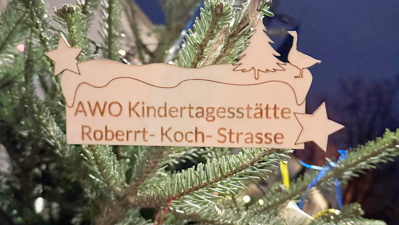 Das Foto zeigt einen festlich geschmückten Weihnachtsbaum. Im Vordergrund hängt ein Holzanhänger in Form eines Sterns und eines Weihnachtsbaums mit der Aufschrift „AWO Kindertagesstätte Robert-Koch-Strasse“. Im Hintergrund sind weitere Dekorationen und Lichter zu sehen, die eine gemütliche Weihnachtsatmosphäre schaffen.