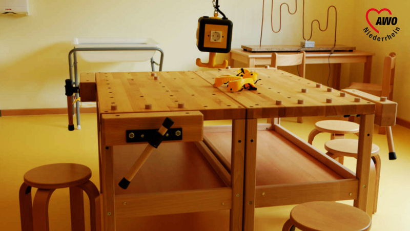 Auf dem Foto ist ein Kita-Raum mit Holzmöbeln zu sehen. Es gibt einen großen Tisch mit einigen Gegenständen darauf, Hocker um ihn herum 