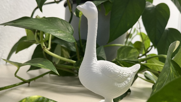 Auf dem Bild steht eine weiße Gänsefigur zwischen üppigen grünen Zimmerpflanzen auf einer hellen Fensterbank. Sie hat einen langen, aufrechten Hals, einen leicht nach links gerichteten Kopf und eine strukturierte Oberfläche, die Federn nachbildet.