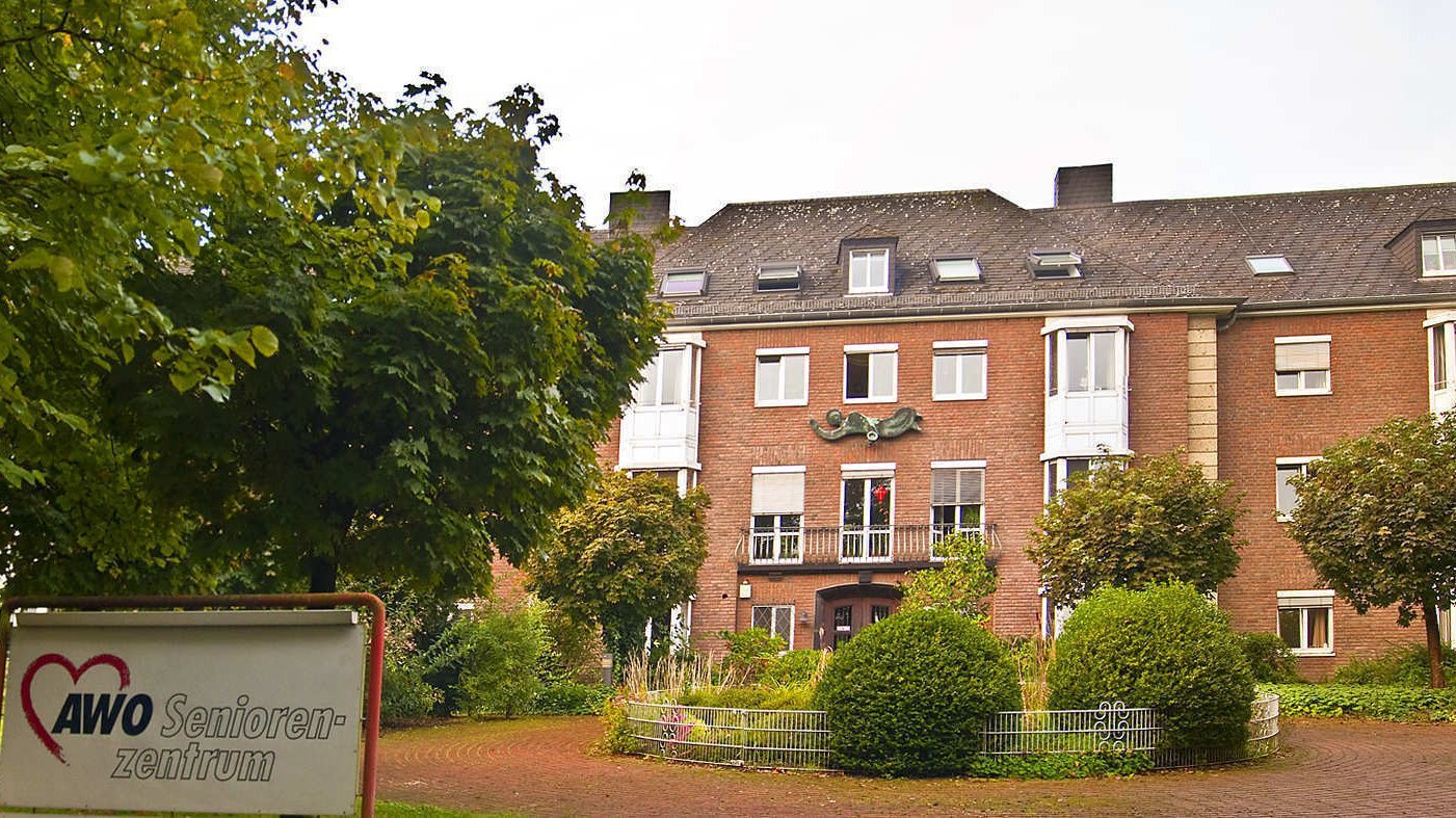 Das Foto zeigt ein Gebäude, das zu einem Seniorenheim gehört, mit dem Schild "AWO Seniorenzentrum" im Vordergrund. Das Gebäude ist aus rotem Backstein, hat mehrere Etagen und ist von Bäumen und Grünanlagen umgeben. Im Vordergrund sieht man einen gepflasterten Bereich mit einem Zaun und einer gepflegten Gartenlandschaft.