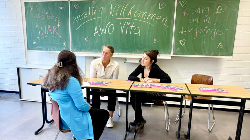 Auf dem Foto sind drei Personen an einem Tisch in einem Klassenraum zu sehen. An der grünen Tafel hinter ihnen stehen verschiedene Sätze, wie z. B. „Projekt INAR“, „Herzlich Willkommen AWO Vita“ und „Komm an in der Pflege“. Auf den Tischen liegen Informationsmaterialien.