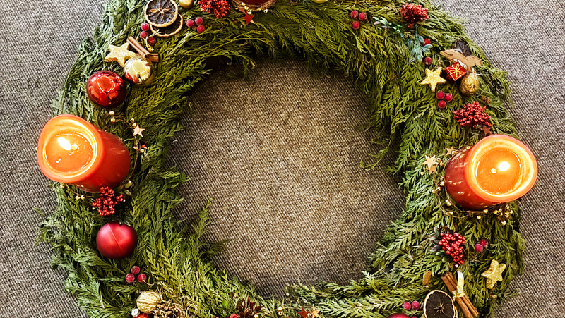 Auf dem Foto ist ein wunderschön geschmückter Adventskranz zu sehen. Der Kranz besteht aus grünen Tannenzweigen und ist mit roten und goldenen Kugeln, getrockneten Orangenscheiben, Zimtstangen, kleinen Sternen und roten Beeren dekoriert. Vier rote Kerzen sind darauf platziert, von denen drei noch nicht angezündet sind und eine bereits brennt. Der Kranz liegt auf einer grauen Oberfläche.