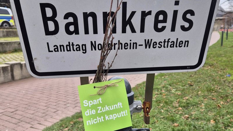 Das Foto zeigt ein Schild, mit den Worten "Bankkreis" und "Landtag Nordrhein-Westfalen". Vor dem Schild ist ein grüner Anhänger befestigt, auf dem steht: "Spart die Zukunft nicht kaputt!". Daneben ist ein Bündel kleiner Zweige angebracht.