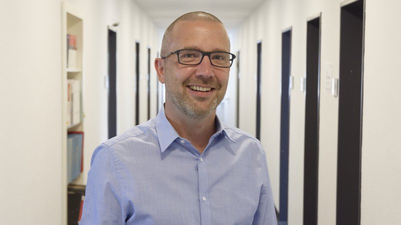 Das Bild zeigt einen Mann mit Brille, der ein hellblaues Hemd trägt und freundlich in die Kamera lächelt. Er steht in einem modernen Büroflur mit mehreren Türen auf beiden Seiten. Links im Bild ist ein Regal mit Ordnern und Büchern zu sehen. Die Atmosphäre wirkt professionell und freundlich.