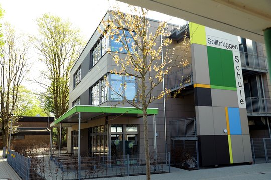 Das Foto zeigt ein modernes Schulgebäude mit der Aufschrift "Sollbrüggenschule" an der Fassade. Das Gebäude ist in neutralen Farbtönen gehalten, und eine markante farbliche Gestaltung an der Seite des Gebäudes ist sichtbar, die sich aus grünen, gelben, blauen und grauen Rechtecken zusammensetzt. Vor dem Gebäude befinden sich ein Baum und ein Zaun. Das Gebäude hat mehrere Stockwerke mit großen Fenstern, und es hat eine überdachte Eingangszone.