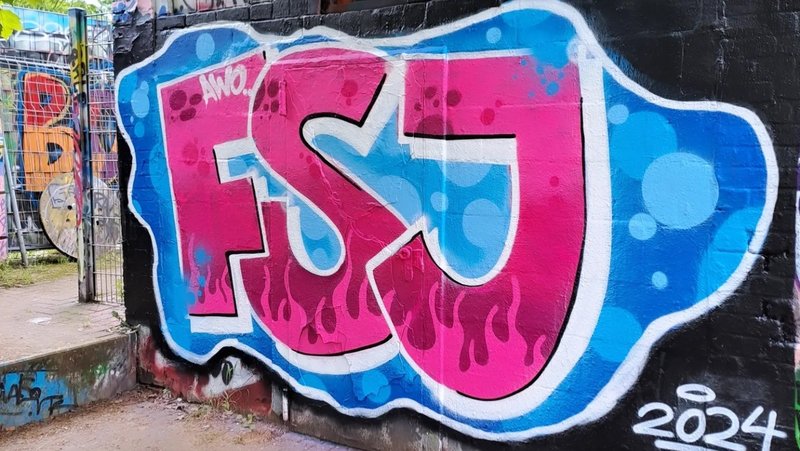 Das Foto zeigt ein Graffiti auf einer Wand. Das Graffiti besteht aus stilisierten Buchstaben, die “FSJ” in großen, blasenartigen Zeichen mit einem 3D-Effekt buchstabieren. Die Buchstaben sind hauptsächlich in Blau- und Pinktönen gemalt, mit weißen Highlights, die den Eindruck von Glanz oder Reflexion erwecken. Unter den Buchstaben befindet sich eine kleinere Inschrift, die “2024” lautet. Die Hintergrundwand ist mit verschiedenen anderen Graffiti-Tags und Markierungen bedeckt, was eine vielschichtige Textur von Straßenkunst erzeugt.