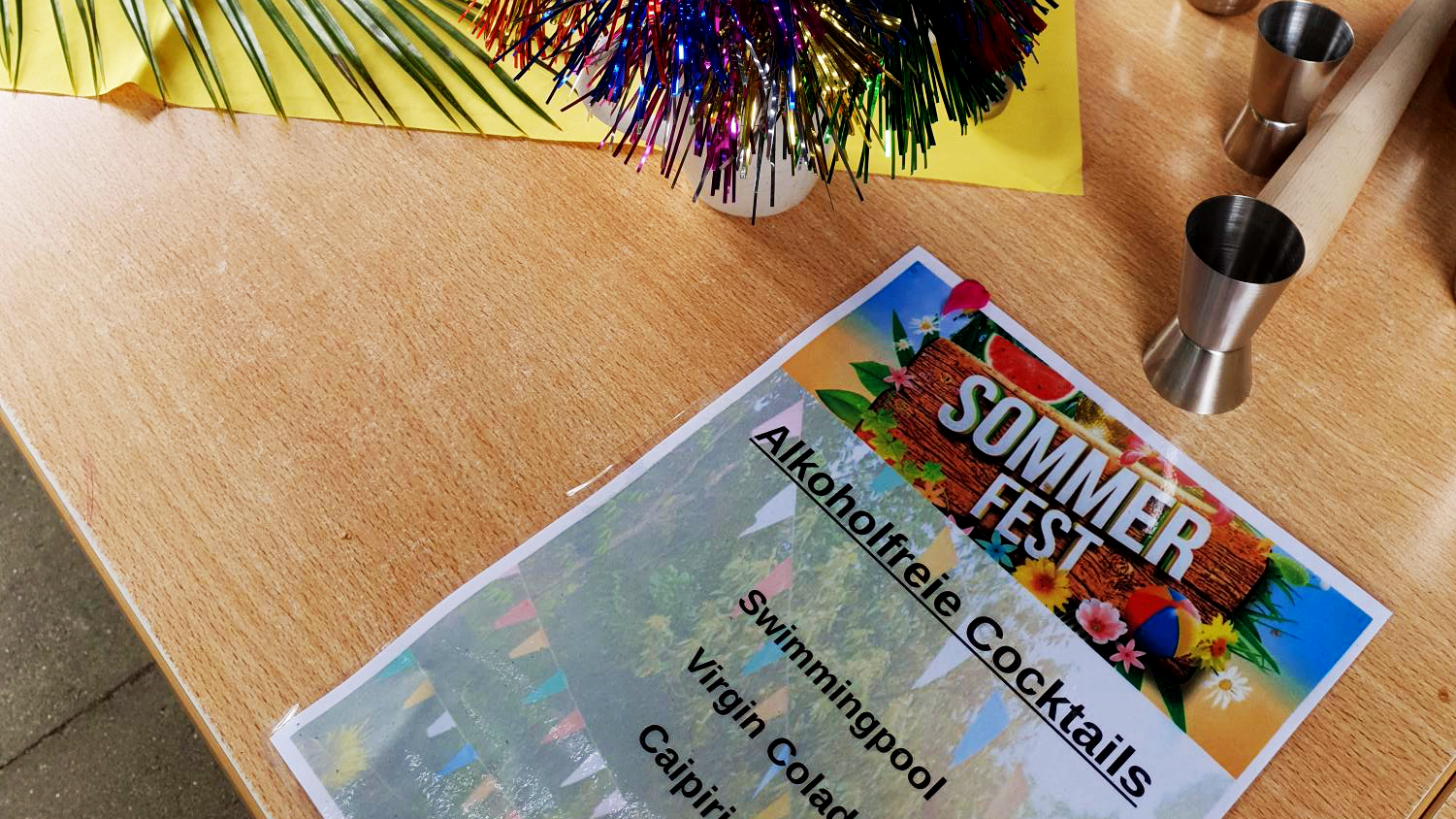 Auf dem Foto ist ein Flyer zum Sommerfest zu sehen. Der Flyer bewirbt alkoholfreie Cocktails wie “Swimmingpool,” “Virgin Colada,” und “Caipirinha.” Außerdem sind Cocktailzubehör und bunte Partydekorationen wie ein gelber Fächer und mehrfarbige Girlanden zu sehen.