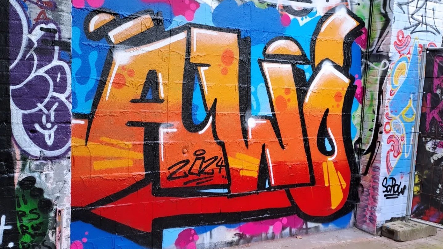 Das Foto zeigt ein Graffiti-Kunstwerk an einer Wand. Das Hauptmerkmal des Graffitis ist das Wort “AWO”, das in großen, stilisierten Buchstaben geschrieben ist. Die Buchstaben sind hauptsächlich orange mit einer weißen Umrandung und Schatteneffekten, was ihnen ein dreidimensionales Aussehen verleiht. Der Hintergrund ist mit verschiedenen anderen Graffiti-Tags und Designs in mehreren Farben gefüllt, was eine lebendige und belebte Szene schafft. 