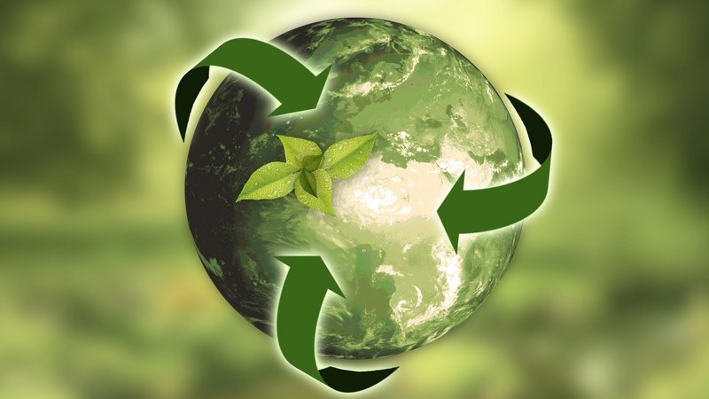 Das Foto zeigt eine grafische Darstellung der Erde mit drei grünen Pfeilen, die ein dreieckiges Recycling-Symbol um sie herum bilden. In der Mitte des Recycling-Symbols befindet sich ein einzelnes grünes Blatt, das scheinbar auf dem Planeten liegt. Der Hintergrund ist verschwommen und in verschiedenen Grüntönen gehalten, was auf Vegetation oder eine natürliche Umgebung hindeutet.