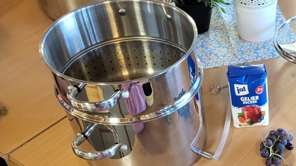 Auf dem Bild sind mehrere Kinder zu sehen, die gemeinsam dunkle Trauben vorbereiten. In der Mitte steht ein großer Edelstahl-Entsafter beziehungsweise Dampfentsafter; daneben liegen weitere Trauben und eine Packung Gelierzucker.
