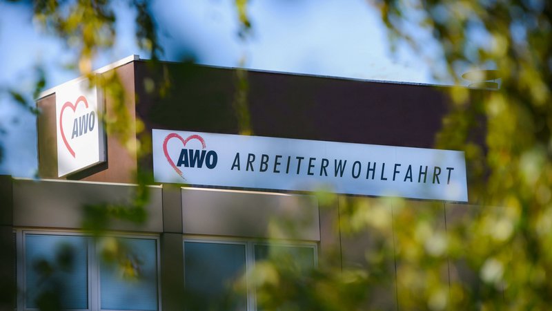 Das Foto zeigt ein Schild mit dem Logo und dem Namen der “Arbeiterwohlfahrt” (AWO), einer deutschen Wohlfahrtsorganisation. Das Schild trägt das AWO-Logo, ein Herz, gefolgt von “AWO” in großen weißen Buchstaben auf rotem Hintergrund, und darunter den vollen Namen “ARBEITERWOHLFAHRT” in schwarzen Buchstaben auf weißem Hintergrund. Das Schild ist auf dem Dach eines Gebäudes montiert. Im Vordergrund sind verschwommene grüne Blätter zu sehen, was darauf hindeutet, dass das Foto durch das Laub hindurch aufgenommen wurde und dem Bild Tiefe und einen natürlichen Rahmen verleiht.