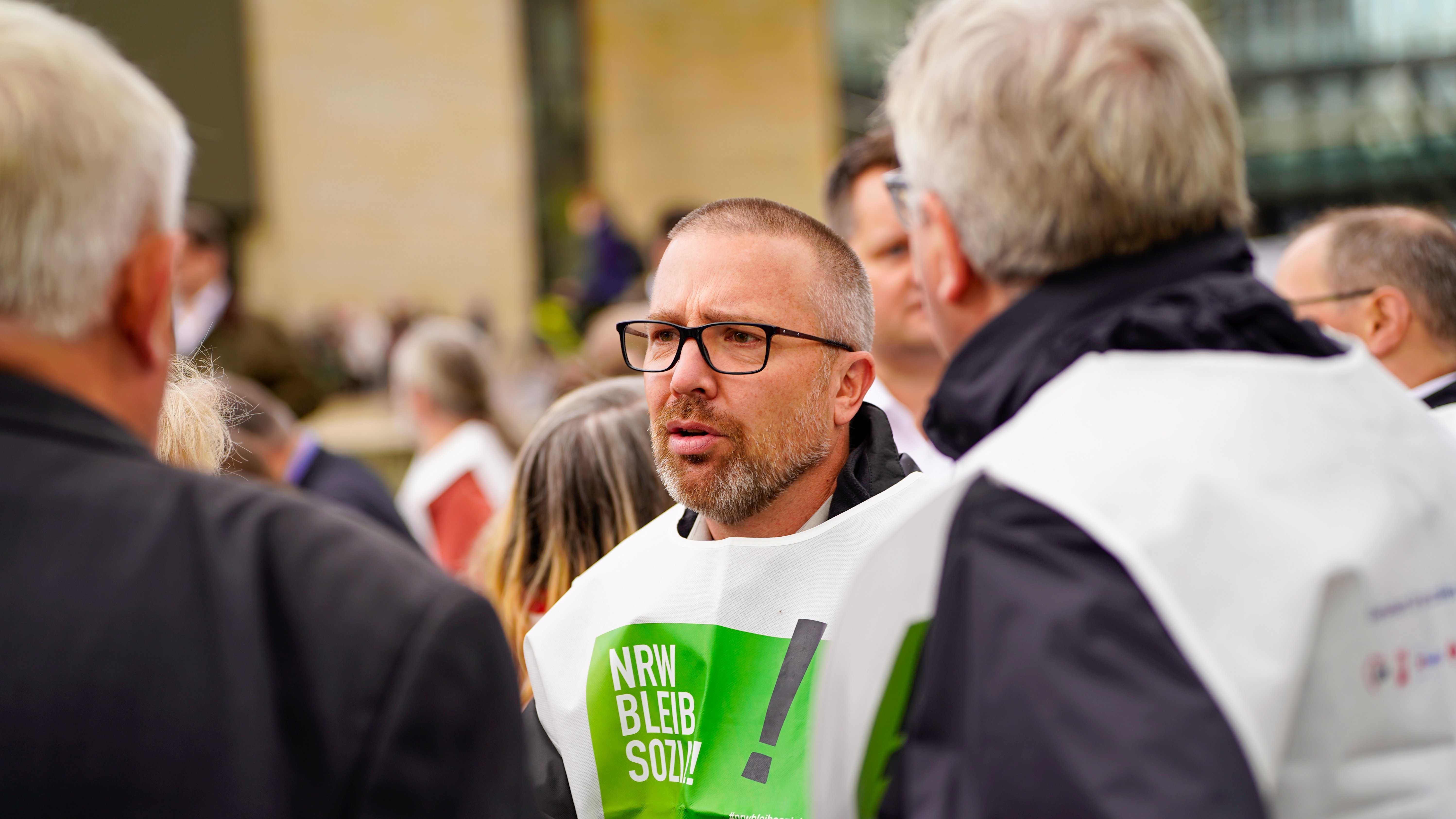 Das Foto zeigt eine Gruppe von Menschen von hinten. Im Vordergrund steht Michael Rosellen, der eine weiße Weste mit grünem Text trägt. Auf der Weste steht “NRW BLEIB SOZIAL" 