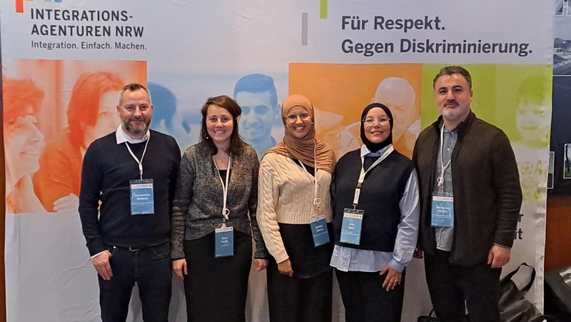 Das Foto zeigt eine Gruppe von fünf Personen, die vor einem Banner der Integrationsagenturen NRW stehen. Das Banner enthält die Slogans "Integration. Einfach. Machen." und "Für Respekt. Gegen Diskriminierung.". Die Personen tragen Namensschilder und sind Teilnehmende des 10. Integrationskongresses des Landes NRW. Im Vordergrund sind ein Tisch mit Broschüren, Unterlagen und Taschen zu sehen, was auf einen Informationsstand oder eine Ausstellung im Rahmen des Kongresses hindeutet. Die Atmosphäre wirkt professionell und engagiert, passend zum Thema Integration und Chancengleichheit.