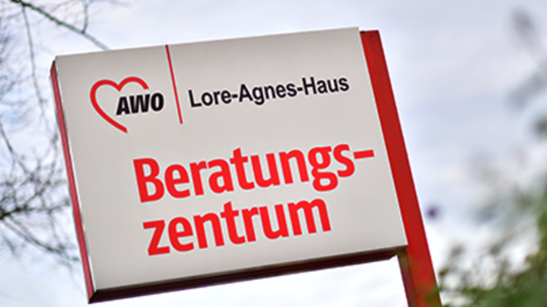 Das Foto zeigt ein Schild mit der Aufschrift: "AWO Lore-Agnes-Haus Beratungszentrum". Das Bild wurde schräg aufgenommen, und das Schild steht vor einem unscharfen Hintergrund mit Bäumen und Himmel.