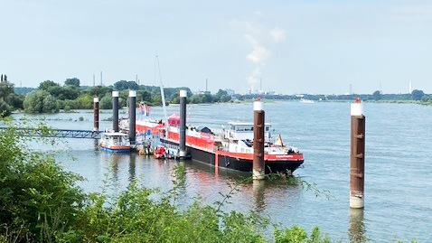 Auf dem Foto ist ein Fluss mit einem rot-weißen Boot zu sehen, das an einem Pier festgemacht ist. Im Wasser befinden sich mehrere vertikale Pfähle zum Festmachen. Im Hintergrund sieht man das andere Ufer mit Bäumen und industriellen Strukturen, darunter eine Fabrik mit Schornsteinen, aus denen Rauch oder Dampf aufsteigt. Der Himmel ist blau mit einigen Wolken.