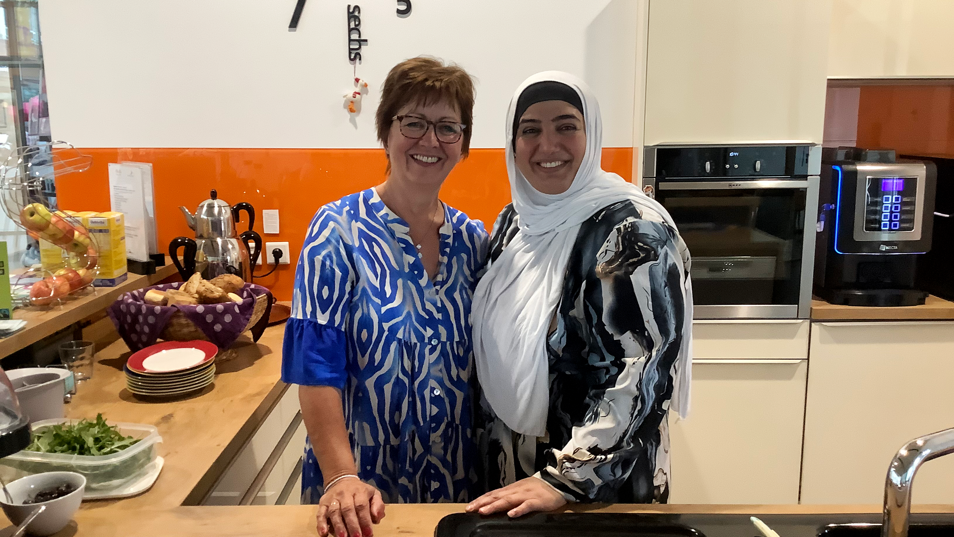 Das Foto zeigt zwei Frauen in einer Cafeteria, die freundlich lächeln. Die Frau auf der linken Seite trägt ein blau-weiß gemustertes Kleid und die Frau rechts einen Hijab und ein schwarz-weiß gemustertes Oberteil. Im Hintergrund befinden sich Küchengeräte wie ein Wasserkocher, ein Kaffeeautomat und einige Speisen, darunter Brot und Obst. Über den beiden Frauen ist eine Wanduhr zu sehen, die moderne Zahlen anzeigt.