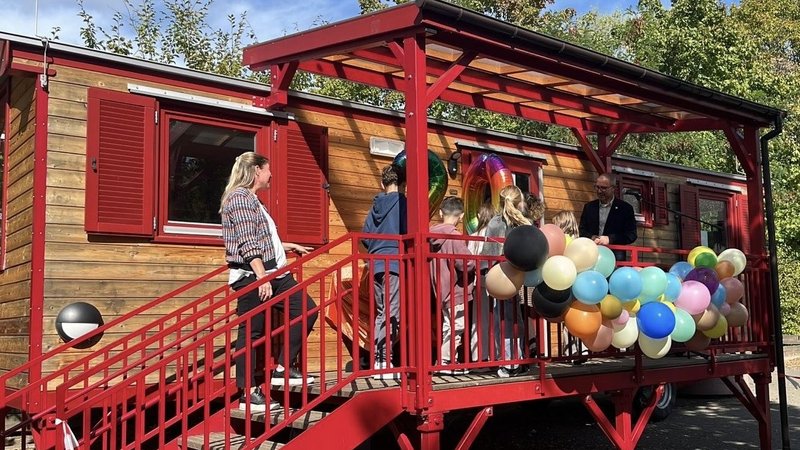 Auf dem Foto ist eine Szene im Freien vor einem Wagen oder einem kleinen, holzverkleideten Gebäude zu sehen. Es scheint sich um eine Art Veranstaltung oder kleine Feier zu handeln, denn: Das Gebäude hat eine auffällige, rot gestrichene Treppe und eine überdachte Veranda. Die Veranda ist mit einem großen Bündel von bunten Ballons (in verschiedenen Pastell- und dunkleren Tönen) geschmückt. Mehrere Personen sind auf der Treppe und der Veranda versammelt, darunter eine erwachsene Frau auf der Treppe, ein erwachsener Mann auf der Veranda und mehrere Kinder. Im Fenster über der Veranda sind auch große, folienartige Zahlen-Ballons zu erkennen, was auf einen Geburtstag oder ein Jubiläum hindeutet. Die Umgebung besteht aus Bäumen und Grünzeug im Hintergrund, und der Himmel ist blau mit Sonnenschein.