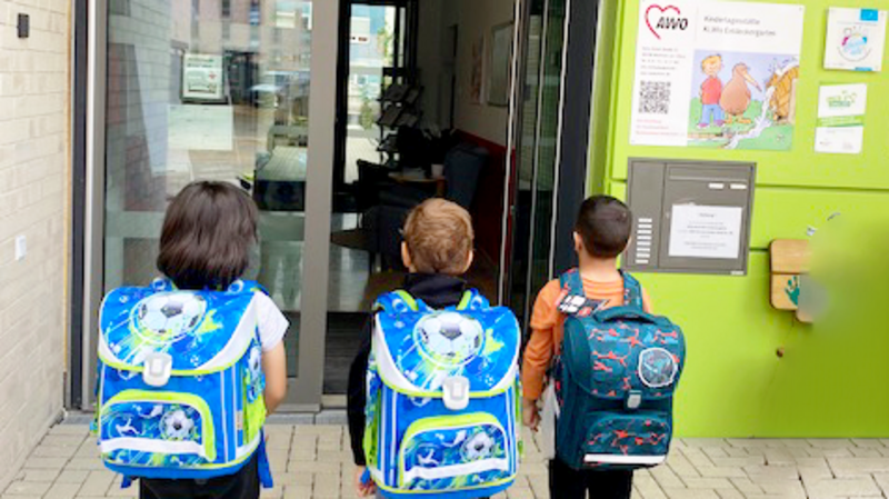 Das Foto zeigt drei Kinder von hinten, die vor einem Glastüreingang zu einem Gebäude stehen. Jedes Kind trägt einen Tornister mit blauen Mustern; die Rucksäcke wirken im Verhältnis zur Größe der Kinder recht groß, was darauf hindeutet, dass sie ziemlich jung sind. Der Eingang des Gebäudes ist mit verschiedenen Plakaten und Schildern versehen.