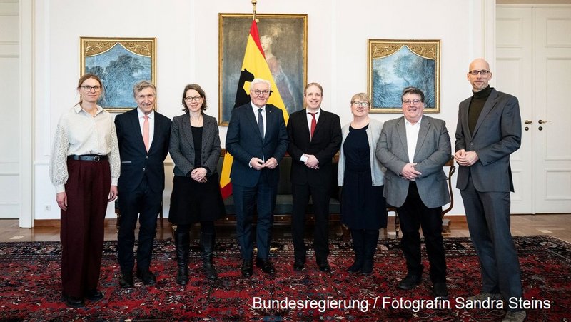 Das Foto zeigt eine Gruppe von acht Personen, die in einem repräsentativen Raum stehen. In der Mitte steht ein Mann mit Brille in einem dunklen Anzug, der Bundespräsident Frank-Walter Steinmeier, vor einer deutschen Flagge. Die Anwesenden sind formell gekleidet und Teil eines Treffens. Im Hintergrund hängen zwei Bilder an der Wand, und der Raum hat einen klassischen Einrichtungsstil mit Teppich und Holzmöbeln. Unten rechts ist eine Quellenangabe: „Bundesregierung / Fotografin Sandra Steins“.