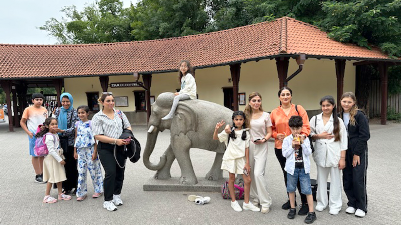 Auf dem Foto ist eine Gruppe von Menschen im Eingangsbereich des Gelsenkirchener Zoos zu sehen, die sich um und auf einer großen Elefantenstatue versammelt haben. Eine Person sitzt auf dem Rücken des Elefanten, während andere in der Nähe stehen.