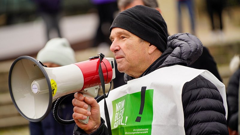 Auf dem Foto ist eine Person zu sehen, die eine weiße Weste mit einem grünen Aufdruck trägt, auf dem "NRW BLEIB SOZIAL!" steht, sowie Hashtags wie #nrwbleibsozial und #kürzungendiemansieht. Die Person hält ein Megafon in der Hand. Sie trägt eine schwarze Mütze und eine dicke schwarze Jacke. Im Hintergrund sind weitere Menschen verschwommen zu erkennen.