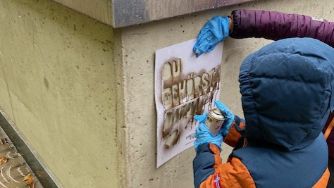 Das Foto zeigt zwei Personen, die eine Schablone an eine Betonwand halten und mit Sprühfarbe besprühen. Die Schablone enthält den Text "DU GEHÖRST DAZU". Die Personen tragen warme Kleidung und Handschuhe. Eine Person hält die Schablone fest, während die andere, ein Kind, die Sprühdose bedient. Auf der Oberseite der Betonwand befindet sich der Deckel der Sprühdose.