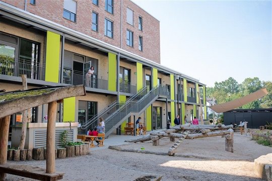 Auf dem Foto ist ein modernes, zweistöckiges Gebäude mit einem großen Außenspielbereich zu sehen. Das Gebäude hat eine Fassade aus Backstein mit grünen Akzenten und große Fenster. Eine Treppe führt vom Erdgeschoss zum oberen Stockwerk, wo Menschen auf einem Balkon stehen. Im Vordergrund gibt es einen Sandspielbereich mit Holzkonstruktionen zum Klettern, Balancieren und Spielen. Mehrere Kinder und Erwachsene sind in der Szene zu sehen – einige sitzen an Picknicktischen, während andere sich unterhalten oder mit den Kindern interagieren. Eine große Sonnensegelstruktur spendet Schatten über einem Teil des Geländes.