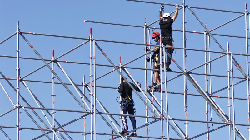 Auf dem Foto sind mehrere Bauarbeiter zu sehen, die an einem Gerüst arbeiten. Sie montieren oder sichern Metallstangen und sind mit Sicherheitsausrüstung ausgestattet, darunter Helme, Gurte und Werkzeuge. Das Gerüst ist hoch und komplex aufgebaut. Im Hintergrund sind ein Gebäude und Bäume zu erkennen, und der Himmel ist klar und blau.