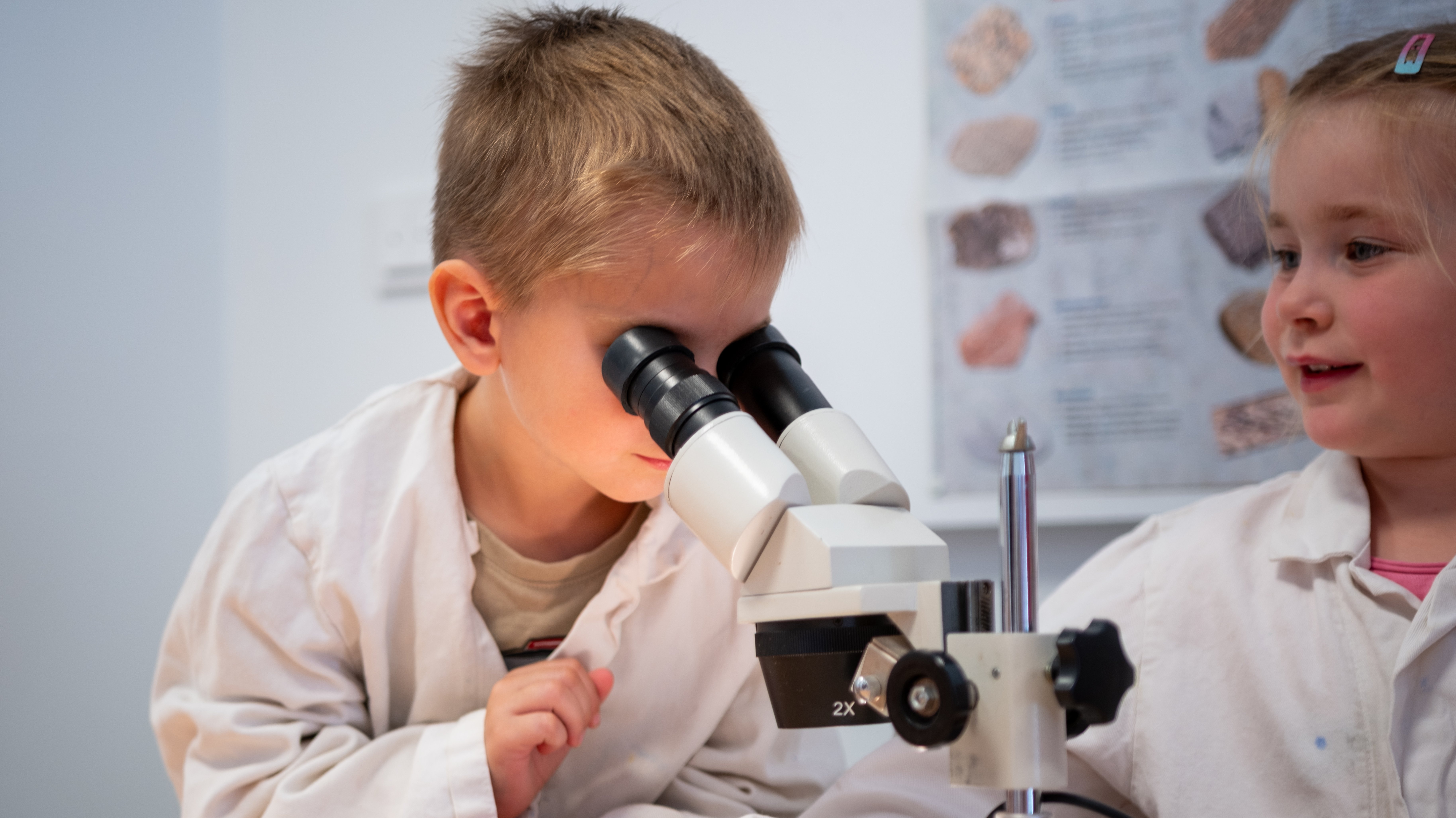 Auf dem Foto sind zwei Kinder zu sehen, die sich mit einem Mikroskop beschäftigen. Der Junge schaut durch das Mikroskop, während das Mädchen daneben sitzt und lächelt. Beide tragen weiße Kittel. Im Hintergrund hängt ein Poster mit Illustrationen, die wie wissenschaftliche oder biologische Abbildungen aussehen.