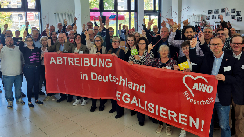 Das Foto zeigt eine Gruppe von Menschen, die in einem hellen Raum stehen. Sie halten einen großen roten Banner, auf dem die Worte „Abtreibung in Deutschland legalisieren!“ zusammen mit dem Logo der AWO (Arbeiterwohlfahrt) Niederrhein abgebildet sind. Einige Personen halten Kleiderbügel hoch, was ein Symbol für die gefährlichen Folgen illegaler Abtreibungen ist. Im Hintergrund sind Fotos an der Wand und Fenster mit Blick nach draußen zu erkennen.