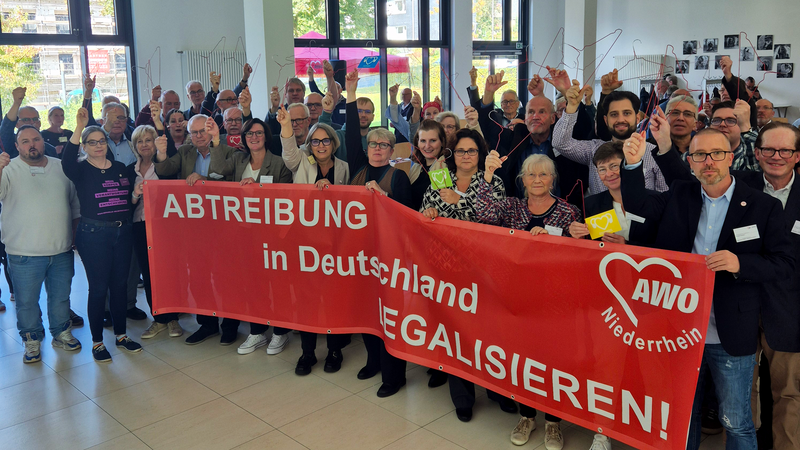 Das Foto zeigt eine Gruppe von Menschen, die in einem hellen Raum stehen. Sie halten einen großen roten Banner, auf dem die Worte „Abtreibung in Deutschland legalisieren!“ zusammen mit dem Logo der AWO (Arbeiterwohlfahrt) Niederrhein abgebildet sind. Einige Personen halten Kleiderbügel hoch, was ein Symbol für die gefährlichen Folgen illegaler Abtreibungen ist. Im Hintergrund sind Fotos an der Wand und Fenster mit Blick nach draußen zu erkennen.