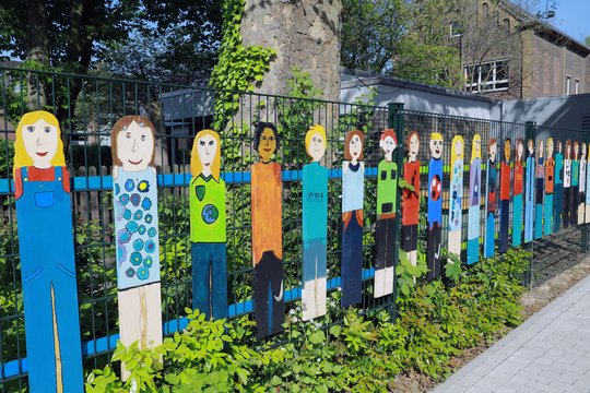 Das Foto zeigt bunte Holzfiguren in einem grünen Beet vor der Schule