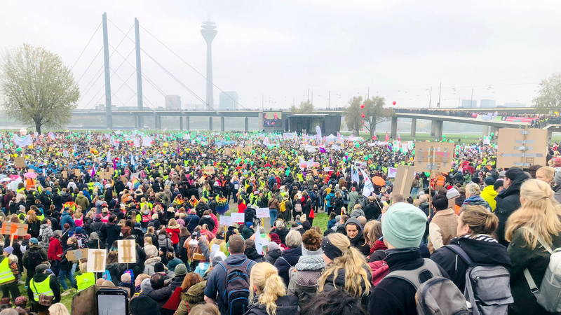 Das Foto zeigt eine große Menschenmenge, die an einer Demonstration oder Kundgebung teilnimmt. Die Menschen tragen bunte Kleidung und halten Schilder und Plakate hoch. Im Hintergrund sind eine Brücke und ein hoher Turm zu sehen. Die Veranstaltung findet im Freien statt, und viele Teilnehmer tragen Rucksäcke und warme Kleidung, was auf kühleres Wetter hinweist. Die Menge ist dicht gedrängt, und einige Personen machen Fotos oder Videos mit ihren Handys.