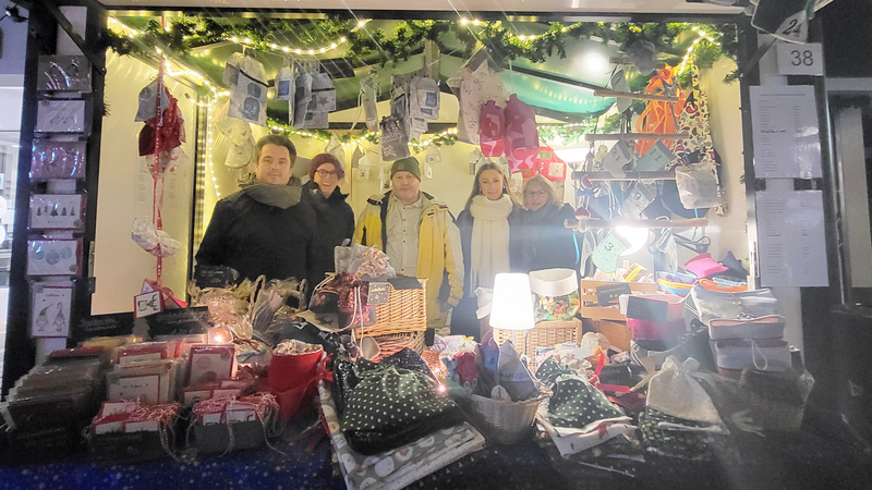 Das Foto zeigt einen Weihnachtsmarktstand mit mehreren Personen, die dahinter stehen. Der Stand ist festlich dekoriert, mit Lichtern, Tannenzweigen und Adventssäckchen, die über den Stand hängen. Auf dem Tisch vor dem Stand werden verschiedene handgefertigte Produkte wie Taschen, Karten, Geschenke und andere dekorative Artikel präsentiert. 