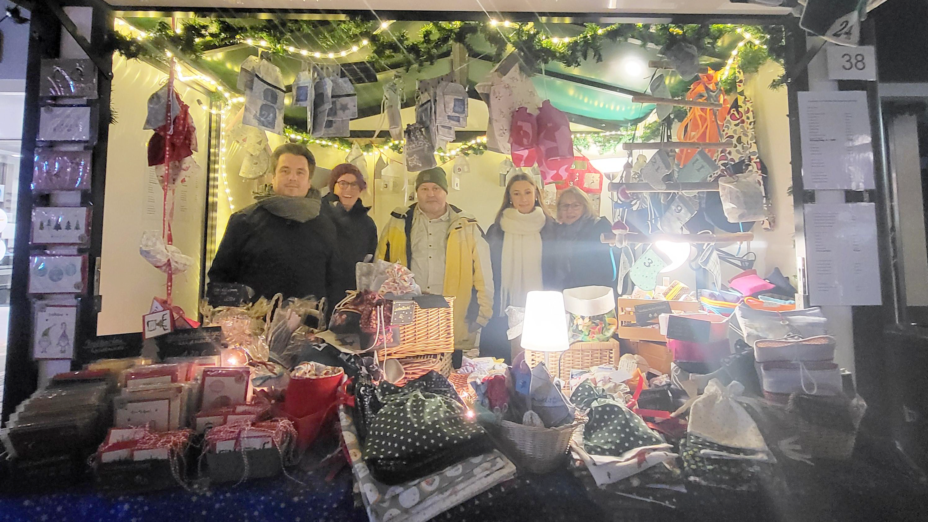 Das Foto zeigt einen Weihnachtsmarktstand mit mehreren Personen, die dahinter stehen. Der Stand ist festlich dekoriert, mit Lichtern, Tannenzweigen und Adventssäckchen, die über den Stand hängen. Auf dem Tisch vor dem Stand werden verschiedene handgefertigte Produkte wie Taschen, Karten, Geschenke und andere dekorative Artikel präsentiert. 