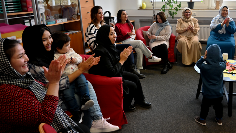 Das Foto zeigt eine Gruppe von Frauen, die in einem Raum auf roten Sesseln und Stühlen sitzen. Sie klatschen und lächeln, was darauf hindeutet, dass sie an einer fröhlichen oder feierlichen Aktivität teilnehmen. Einige Frauen tragen Kopftücher, andere haben offene Haare. In der Mitte des Bildes hält eine Frau ein kleines Kind auf ihrem Schoß, während vorne ein weiteres Kind in einem blauen Kapuzenpullover steht und sich möglicherweise die Ohren zuhält. Im Hintergrund sieht man Regale mit Ordnern und Büchern, sowie Pflanzen und Fenster, die den Raum erhellen. 