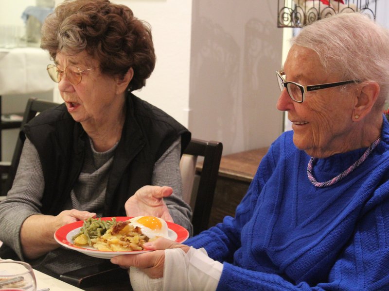 Auf dem Foto reicht eine Bewohnerin einer anderen Bewohnerin den Teller mit dem leckeren Essen.