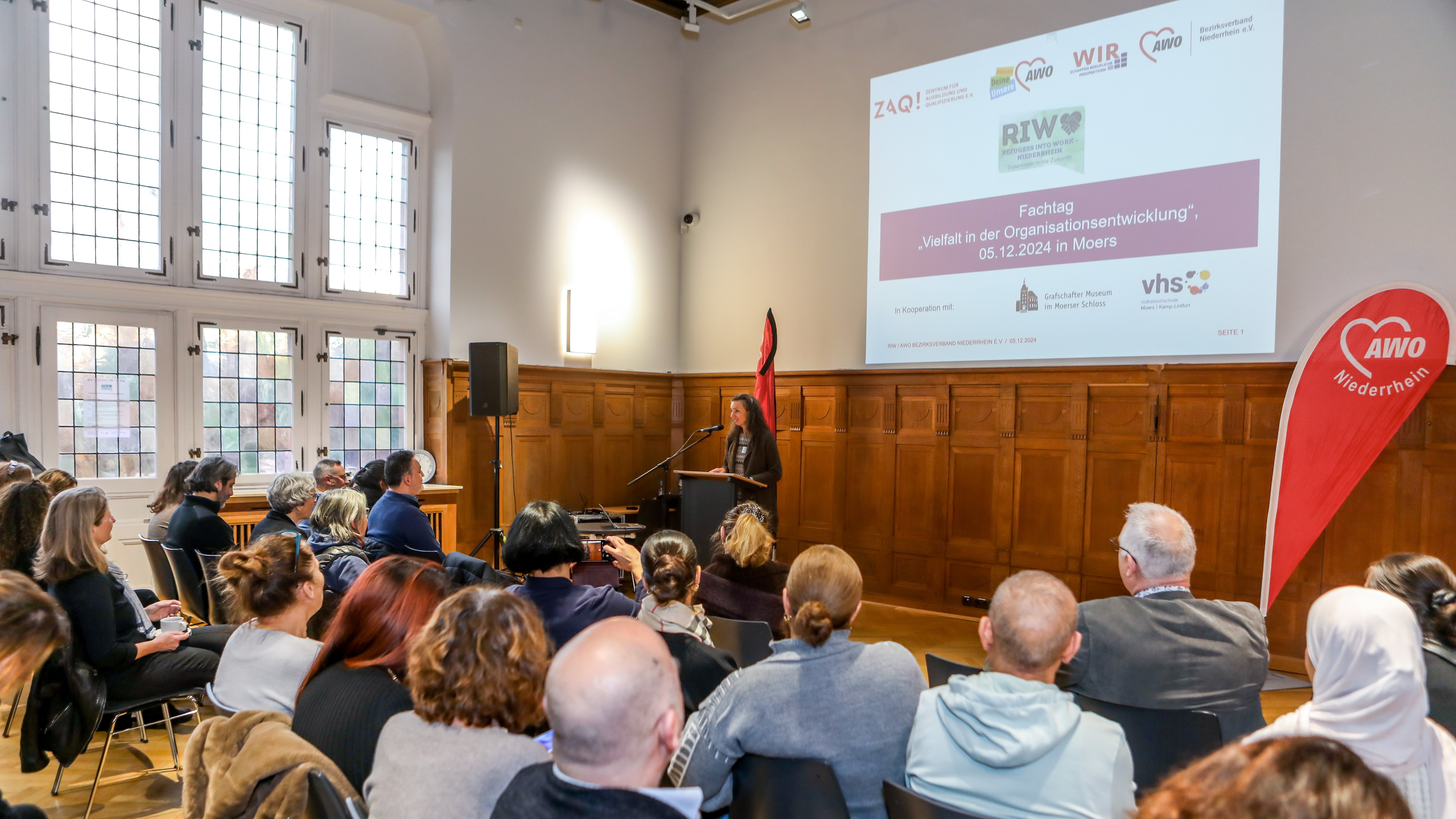 Das Foto zeigt eine Veranstaltung in einem Konferenzraum. Mehrere Personen sitzen auf Stühlen und hören einer Person zu, die an einem Rednerpult steht und eine Präsentation hält. An der Wand hinter der Rednerin ist eine große Leinwand mit einer Projektion zu sehen. Auf der Projektion steht: Fachtag 'Vielfalt in der Organisationsentwicklung' 05.12.2024 in Moers