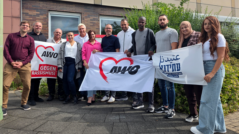 Auf dem Foto ist eine Gruppe von etwa einem Dutzend Personen zu sehen, die vor einem Gebäude stehen. Sie halten drei Banner hoch: Ein weißes Banner mit einem roten Herz und dem Slogan "AWO" in Rot. Ein weiteres weißes Banner mit einem roten Herz und dem Text "AWO GEGEN RASSISMUS!" in Schwarz. Ein weißes Banner mit Text in einer ausländischen Schrift. Die meisten Personen auf dem Foto tragen lässige Kleidung. Die Stimmung wirkt positiv und die Personen schauen freundlich in die Kamera.