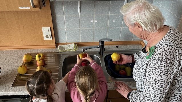 Dieses Bild zeigt Kindergartenkinder wie sie mit einer Bewohnerin das eingekaufte Obst spülen.