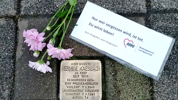 Das Foto zeigt einen sogenannten "Stolperstein," ein in den Boden eingelassenes Gedenksteinpflaster, das an eine Person erinnert, die Opfer des Nationalsozialismus wurde. Auf dem Stolperstein steht: Hier wohnte Hermine Kaesbach Jg. 1917 Seit 1935 in mehreren Heil- und Pflegeanstalten „Verlegt“ 7.3.1941 Landesanstalt Bernburg Ermordet 7.3.1941 Aktion T4 Daneben liegen Blumen und ein Zettel mit der Aufschrift: "Nur wer vergessen wird, ist tot. Du wirst leben!" Darunter steht: "Kein Vergeben, kein Vergessen!" Mit dem Logo der AWO (Arbeiterwohlfahrt) und dem Hinweis auf das Beratungszentrum Lore-Agnes-Haus.