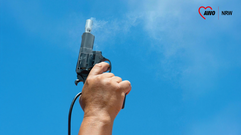 Auf dem Foto ist eine Hand zu sehen, die eine Startpistole in den Himmel hält. Die Pistole scheint gerade abgefeuert worden zu sein, da Rauch aus dem Lauf austritt. Der Hintergrund besteht aus einem klaren blauen Himmel. Außerdem ist oben rechts das Logo der AWO NRW zu sehen. 