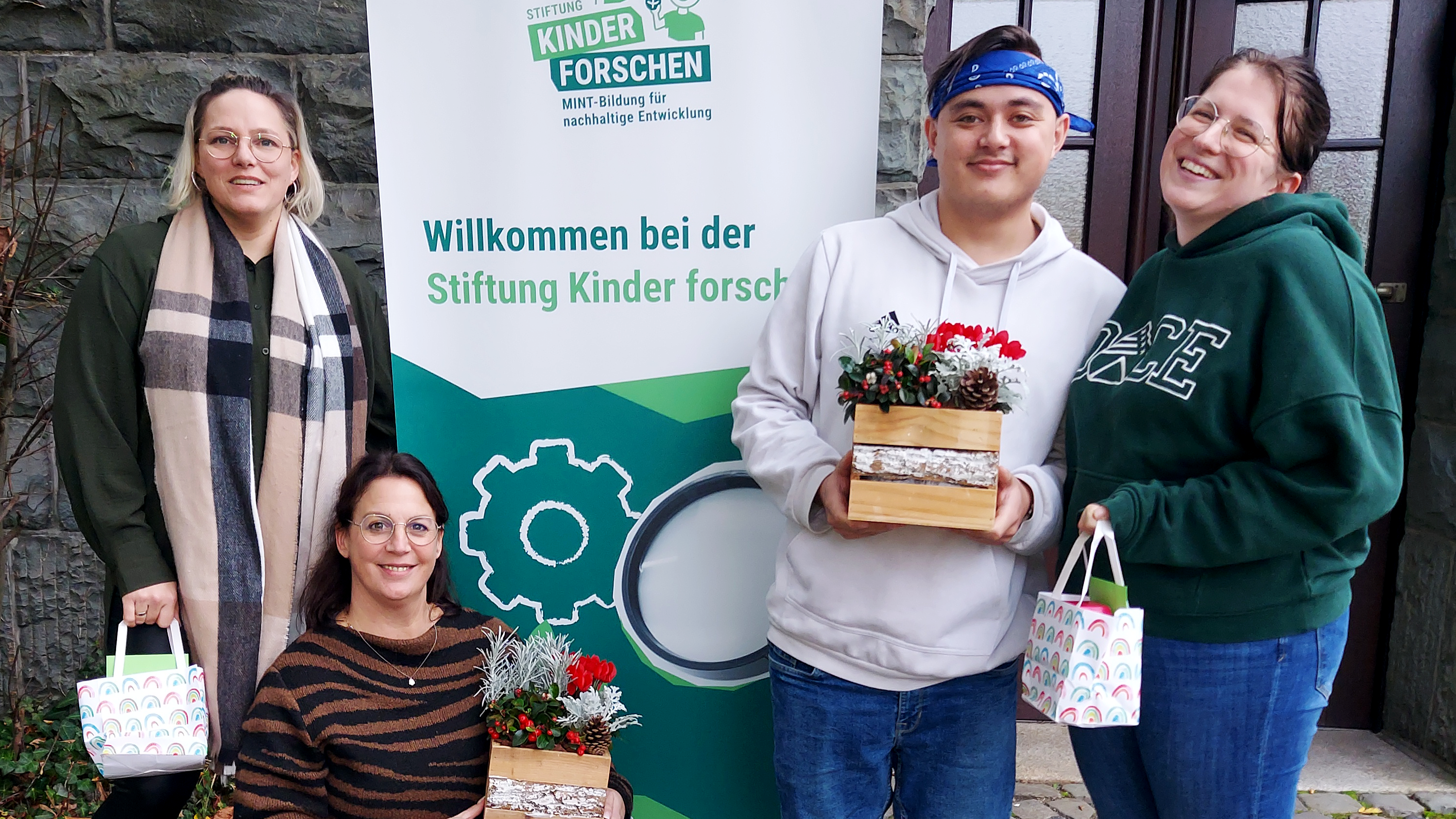 Auf dem Foto ist eine Gruppe von vier Personen zu sehen, die vor einem Banner der "Stiftung Kinder forschen" stehen, welches den Slogan "Willkommen bei der Stiftung Kinder forschen" trägt. Die Personen lächeln und halten kleine Geschenktüten und dekorative Blumengestecke in Holzboxen. 