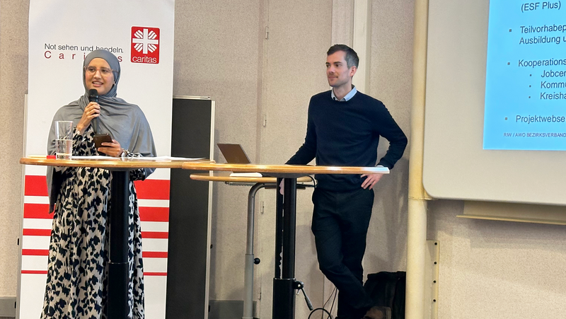 Das Foto zeigt eine Präsentationssituation in einem Konferenzraum. Links steht eine Frau in einem gemusterten Kleid mit einem grauen Kopftuch, die in ein Mikrofon spricht. Rechts daneben steht ein Mann in dunkler Kleidung, der sich an einen Stehtisch lehnt. Im Hintergrund ist ein Banner mit dem Logo von Caritas und der Schrift „Not sehen und handeln. Caritas“ zu erkennen. Rechts im Bild ist eine Projektpräsentation mit dem Titel „ZAQ – Allgemeines“ auf einer Leinwand zu sehen. 