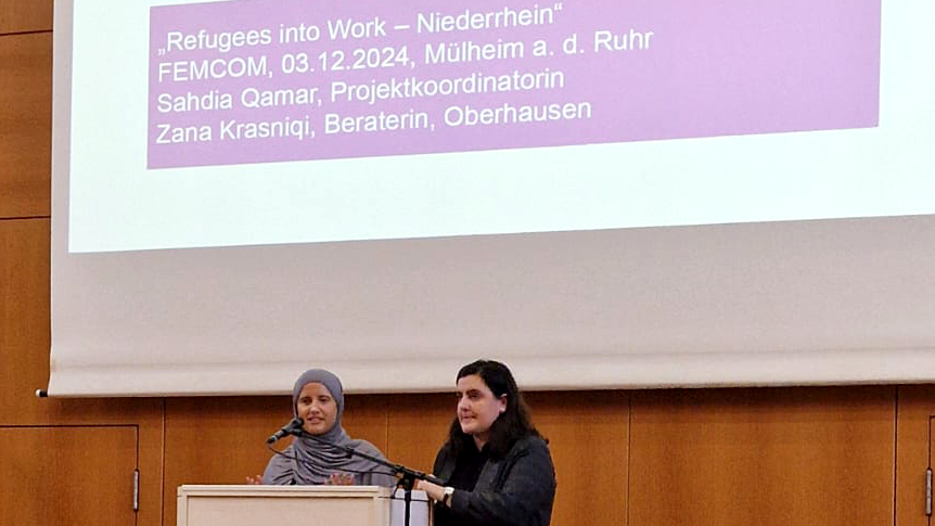 Das Foto zeigt eine Präsentationmit dem Titel „Refugees into Work – Niederrhein“. Die Veranstaltung fand am 03.12.2024 in Mülheim an der Ruhr statt. Auf dem Podium stehen zwei Personen, die eine Präsentation geben. Auf dem Rednerpult ist das Logo von FEMCOM zu sehen. Auf der Leinwand hinter den Personen steht: „Refugees into Work – Niederrhein“ FEMCOM, 03.12.2024, Mülheim a. d. Ruhr Sahdia Qamar, Projektkoordinatorin Zana Krasniqi, Beraterin, Oberhausen