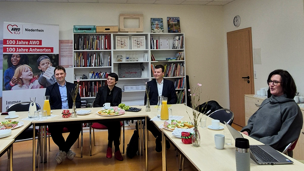 Das Foto zeigt eine Gesprächsrunde in einem Konferenzraum. Zu sehen sind mehrere Personen, die an Tischen sitzen, die in U-Form angeordnet sind. Die Tische sind mit Getränken, kleinen Snacks und Blumen dekoriert. Im Hintergrund befinden sich Bücherregale und ein Banner mit dem Logo der AWO Niederrhein (Arbeiterwohlfahrt) und der Aufschrift „100 Jahre AWO – 100 Jahre Antworten“.