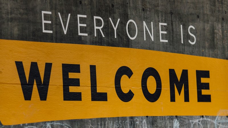 Das Foto zeigt eine Wand mit der Aufschrift „EVERYONE IS WELCOME“ (auf Deutsch: „Jeder ist willkommen“). Die Worte sind in weißer und schwarzer Schrift auf einem gelben Hintergrund geschrieben. Darunter befinden sich Kreidezeichnungen und Kritzeleien, darunter Herzen, Blumen, Gesichter und verschiedene andere Motive auf der grauen Betonwand.